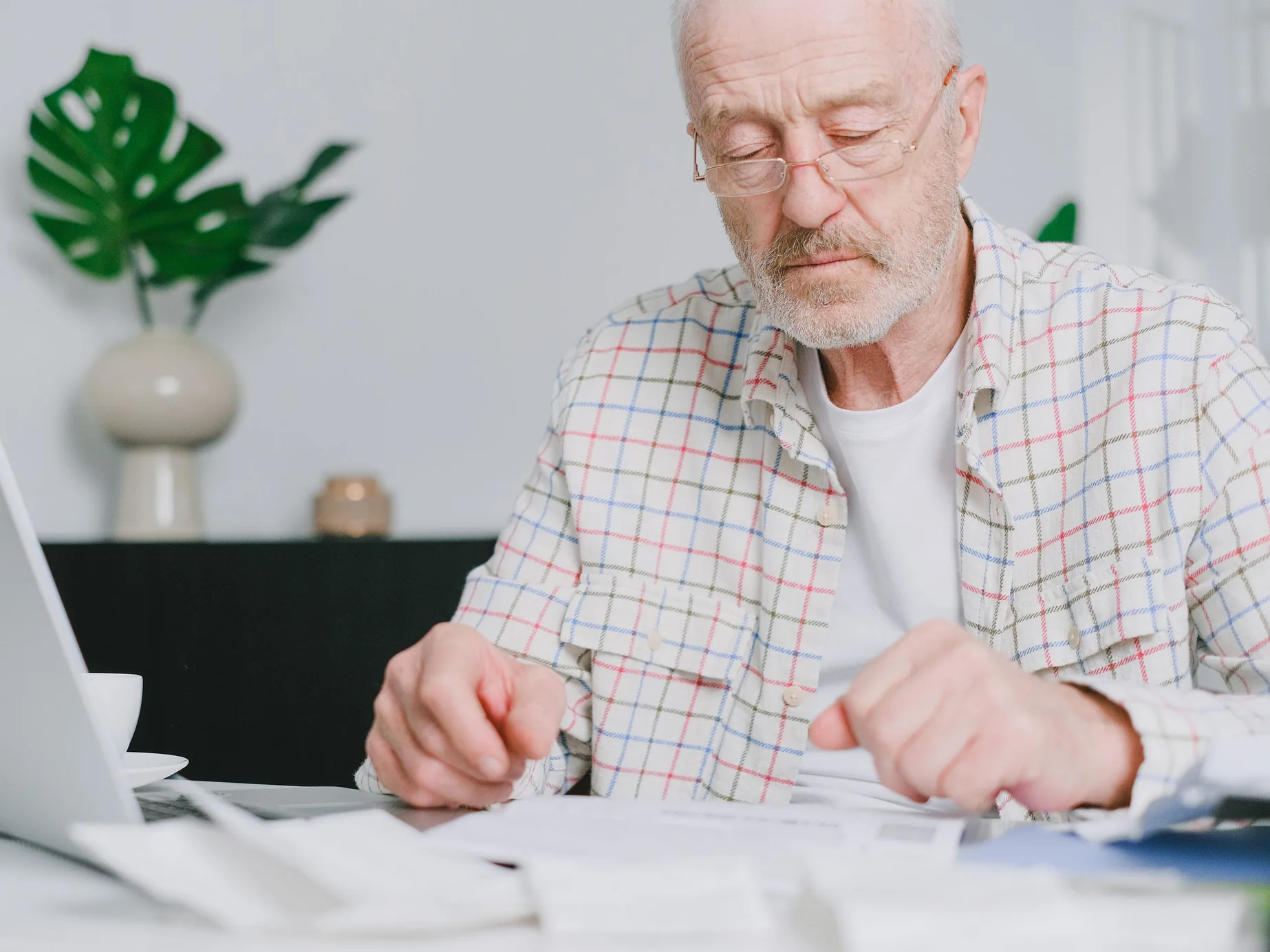 An older man in a checked shirt reads a page closely at his desk, his laptop open beside him