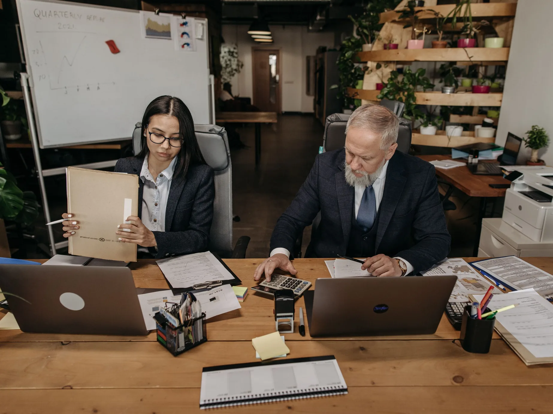 An older owner with a calculator and a younger colleague with a folder work at a shared desk covered in papers and laptops