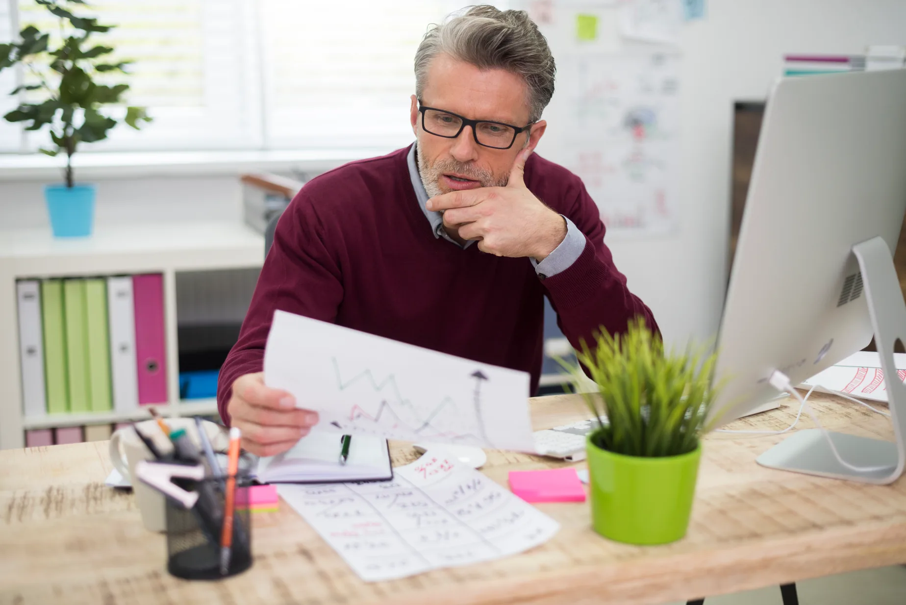 An owner in glasses and a burgundy sweater frowns at a printed chart at his desk, his hand at his chin