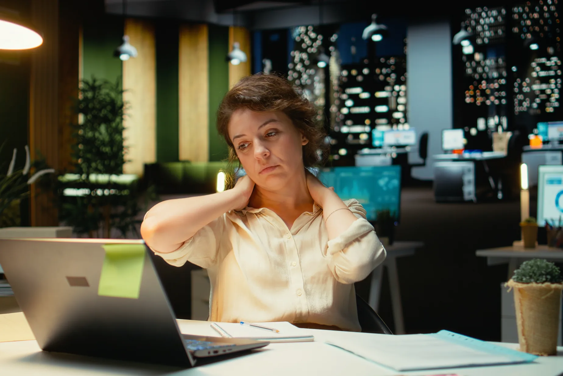 An owner rubs her neck at her laptop late in the evening, the office dark behind her