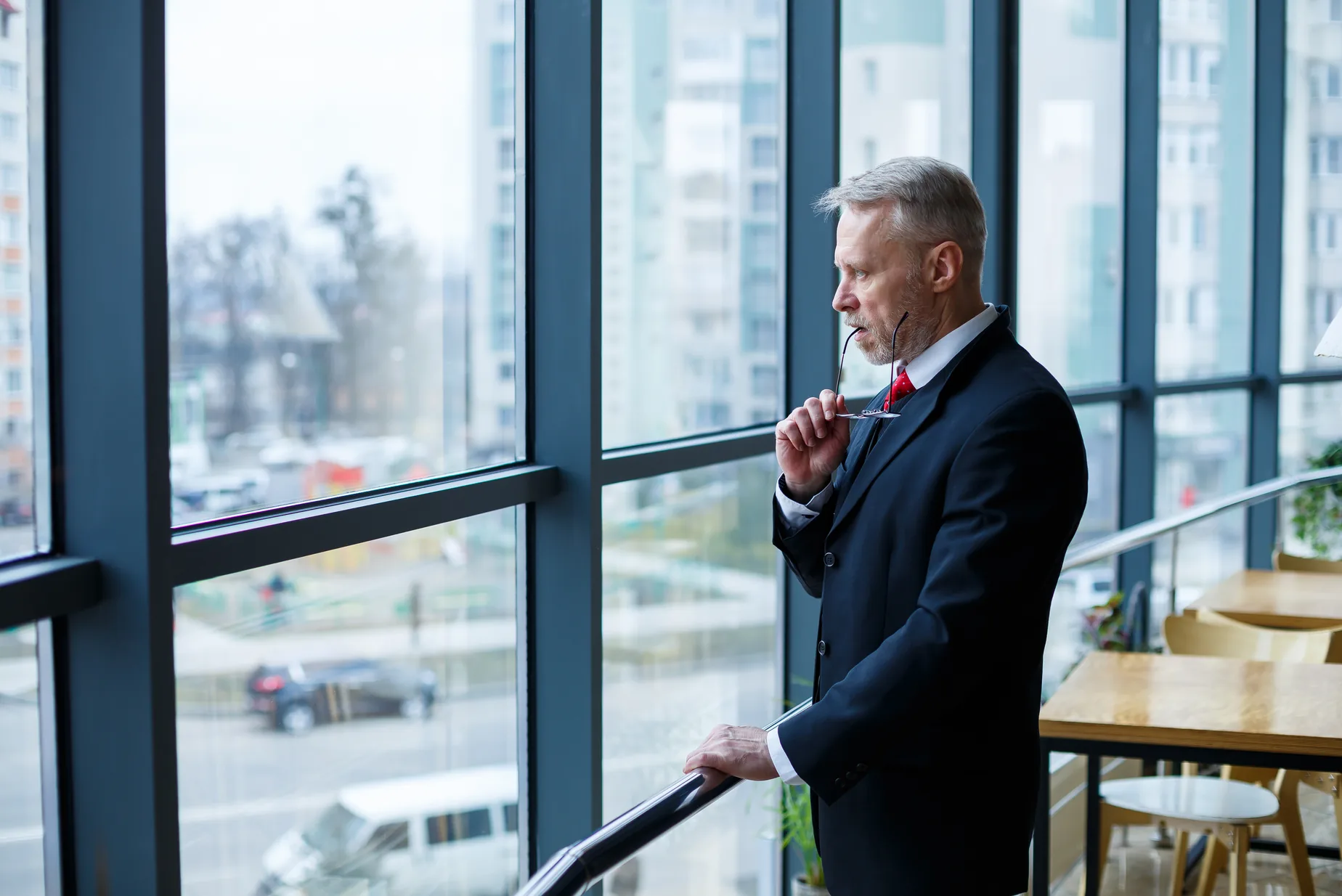 An owner in a dark suit stands at a tall office window, looking out, his glasses in his hand