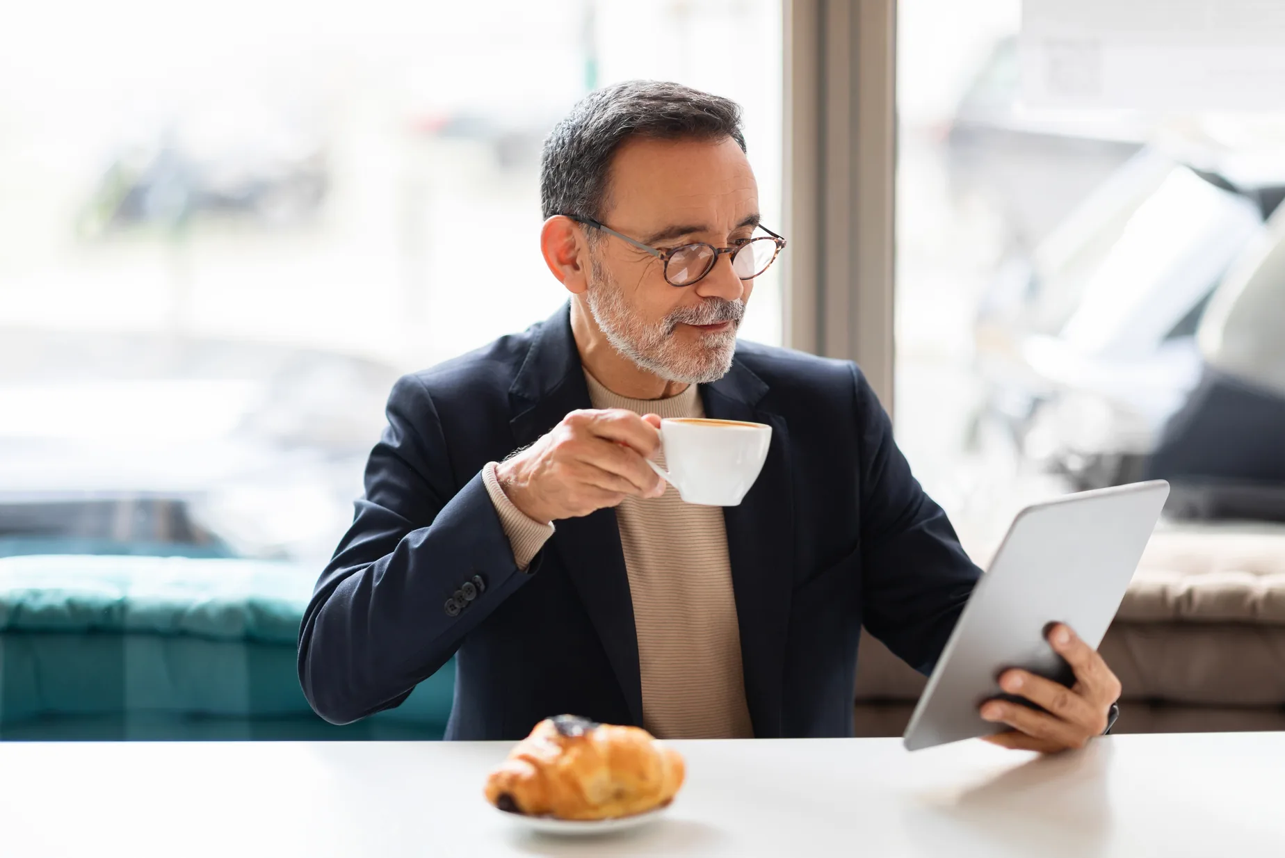 An owner in glasses and a blazer reads on a tablet with a coffee in his hand