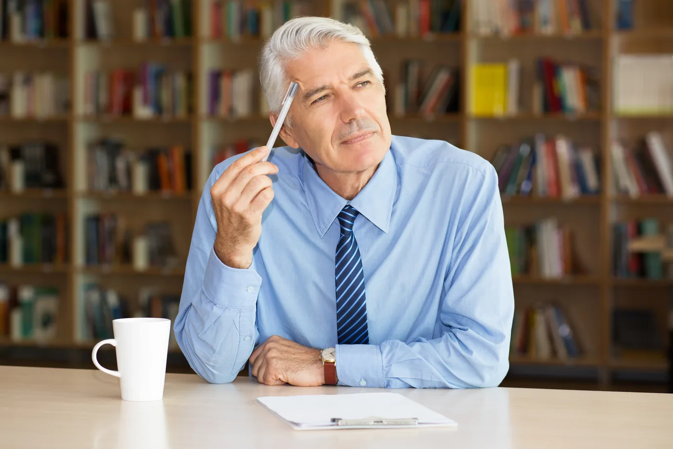 A gray haired owner in a shirt and tie sits at his desk with a pen raised to his temple, thinking, bookshelves behind him
