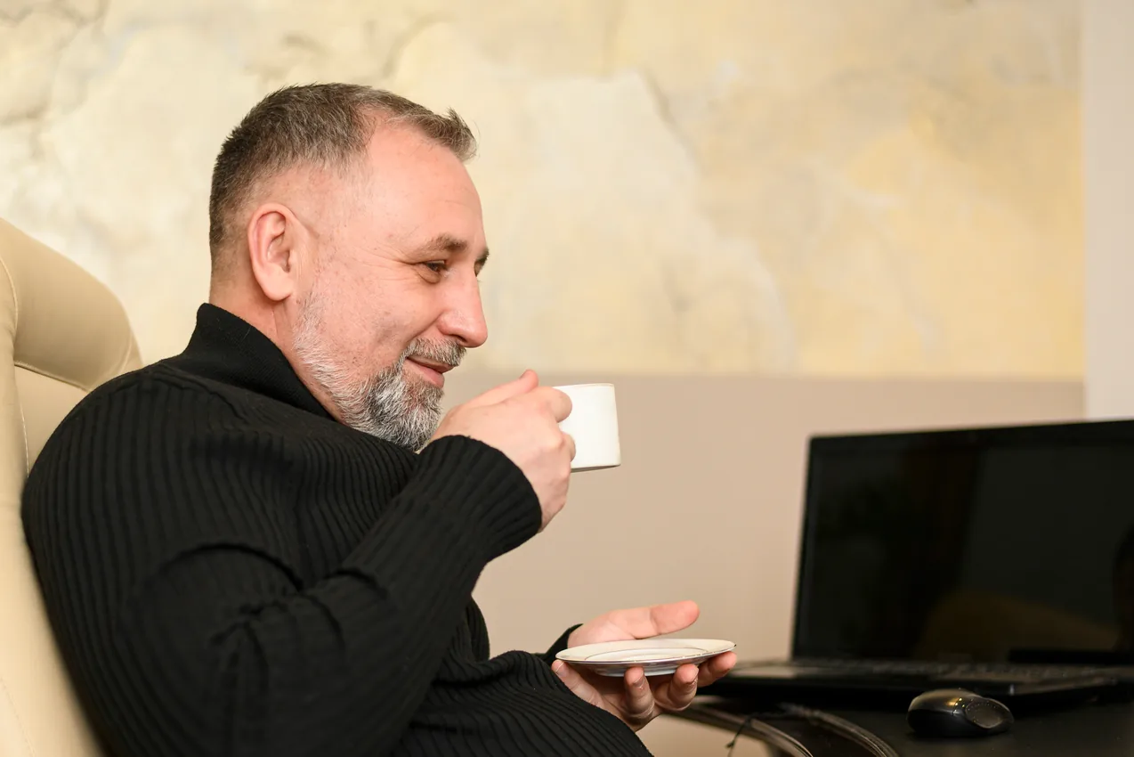 A gray bearded owner in a black sweater smiles as he sips coffee in his office, a laptop open beside him
