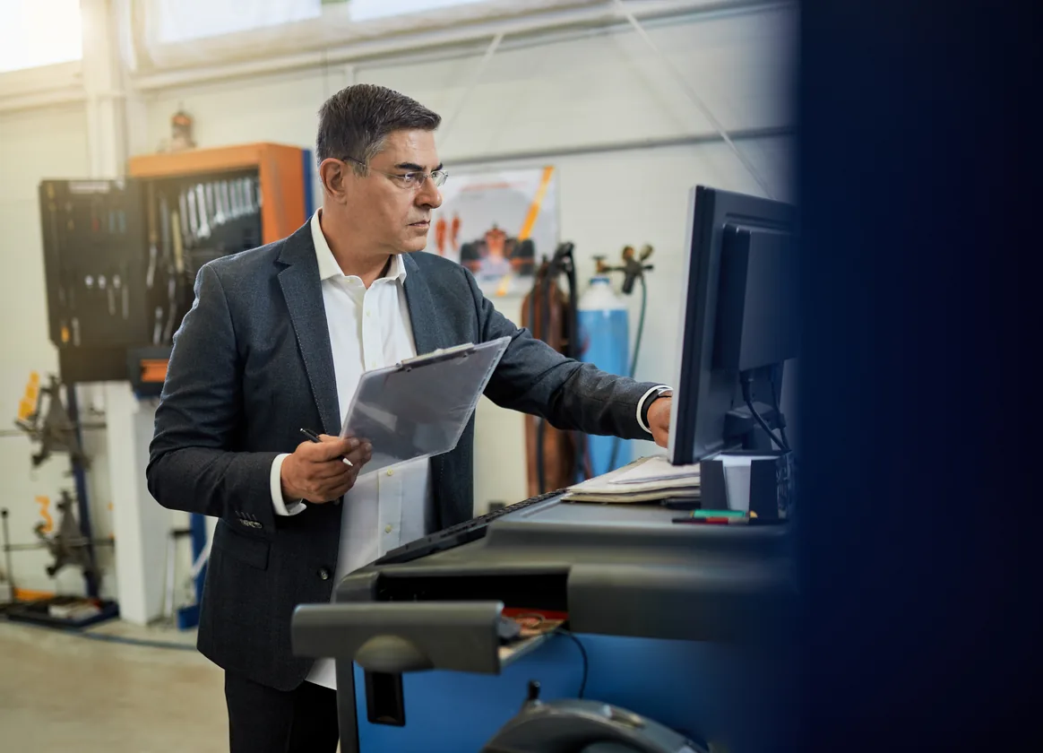 An owner in a dark suit holds a clipboard and checks a screen in his own repair workshop, tools on the wall behind him