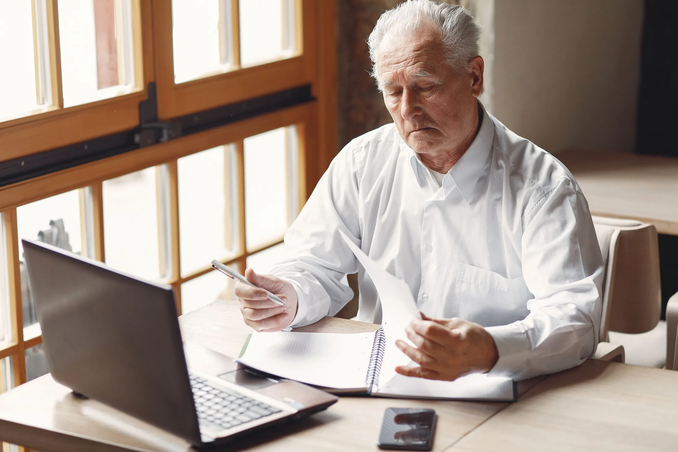 An older man in a white shirt sits at a desk by a sunny window, going through papers with a pen in his hand