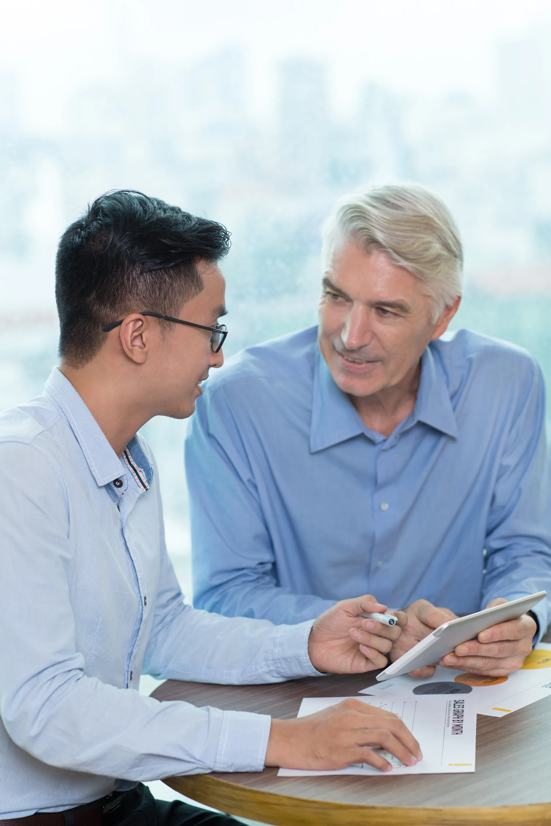 A senior owner with gray hair and a younger colleague in glasses sit at a round table by a window, going over figures on a tablet and printed charts