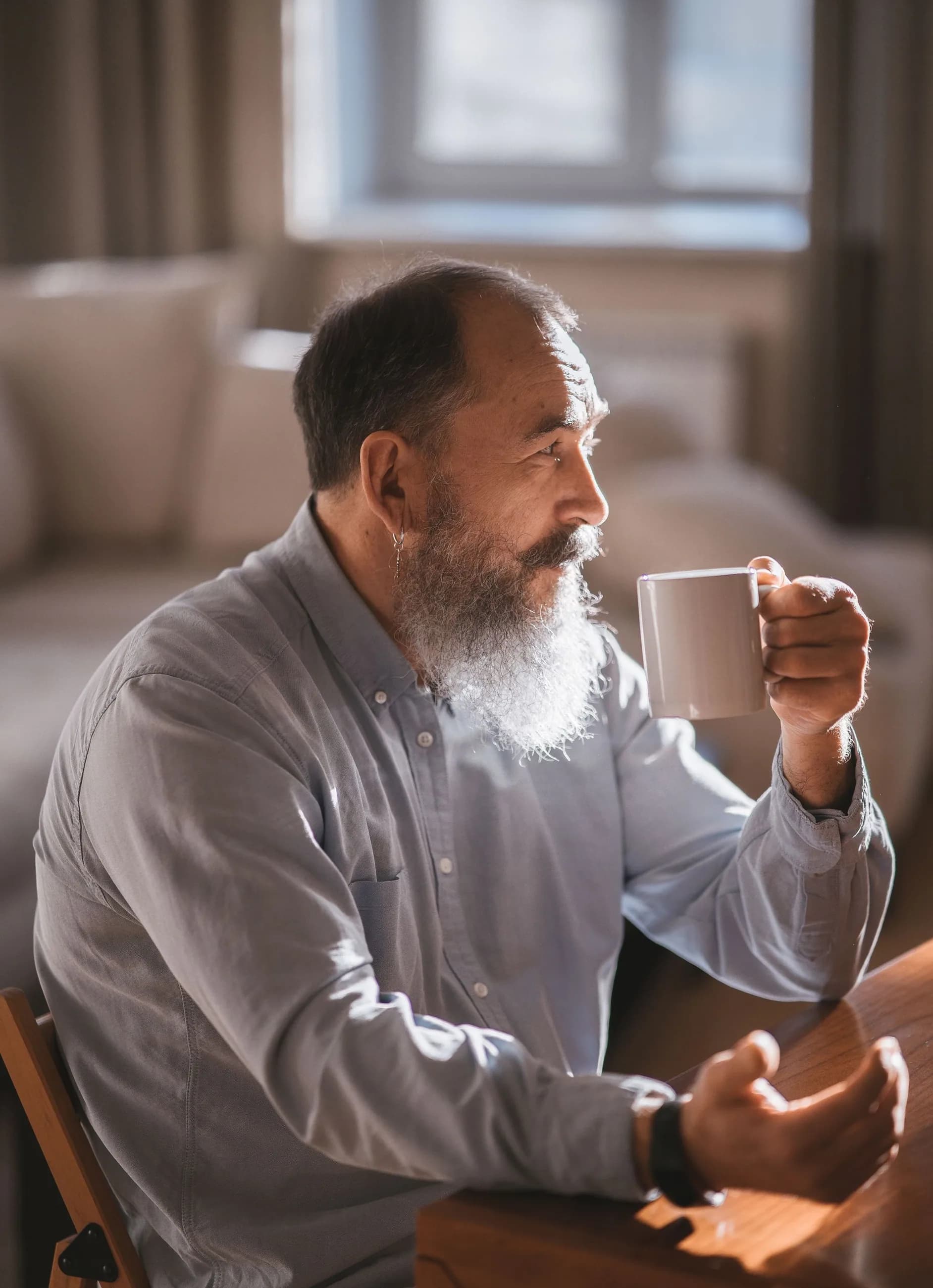 A gray bearded man holds his morning coffee at a wooden table at home, looking out in soft morning light