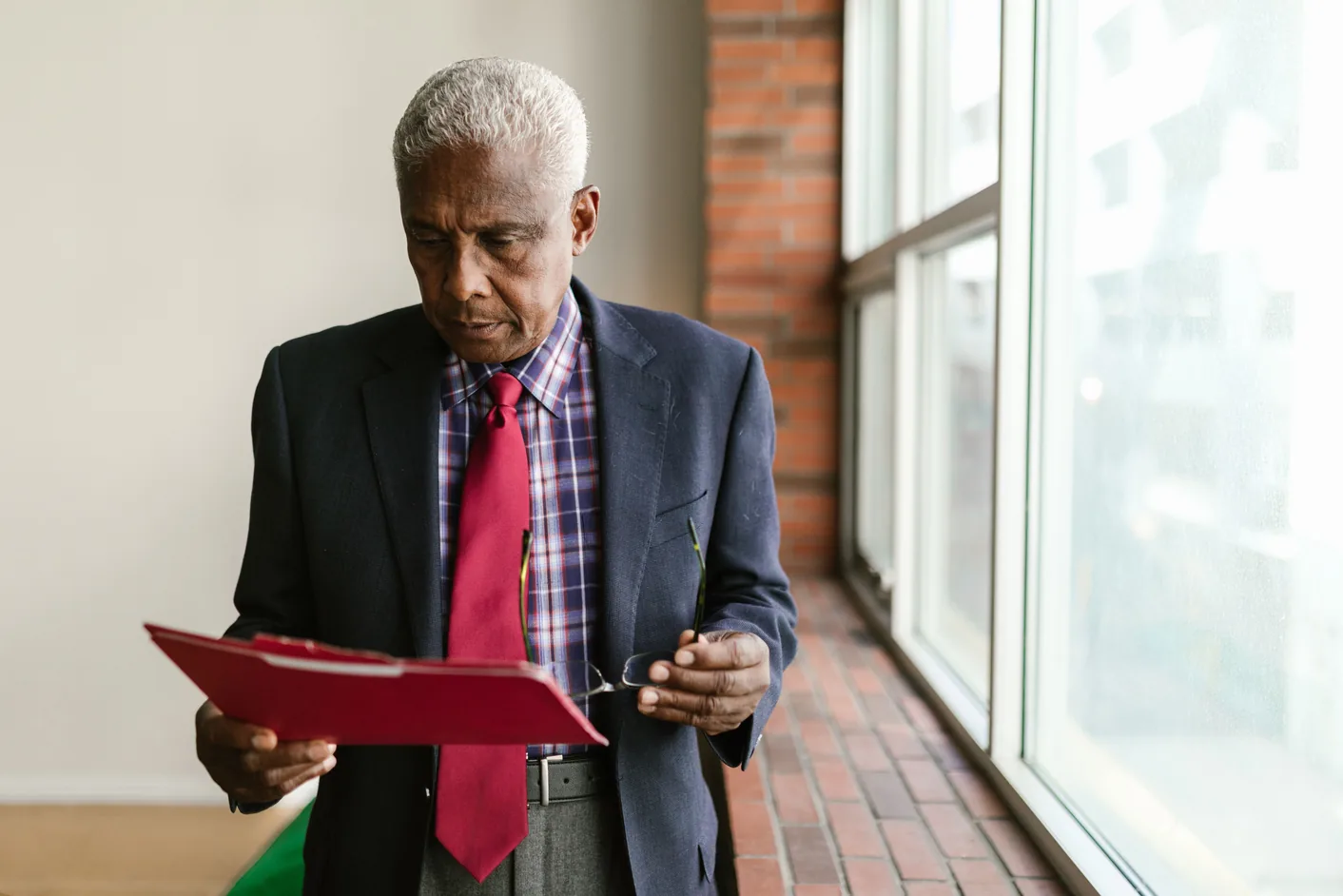 An owner in a dark jacket and red tie stands by a tall window reading a report in a red folder, his glasses in his hand